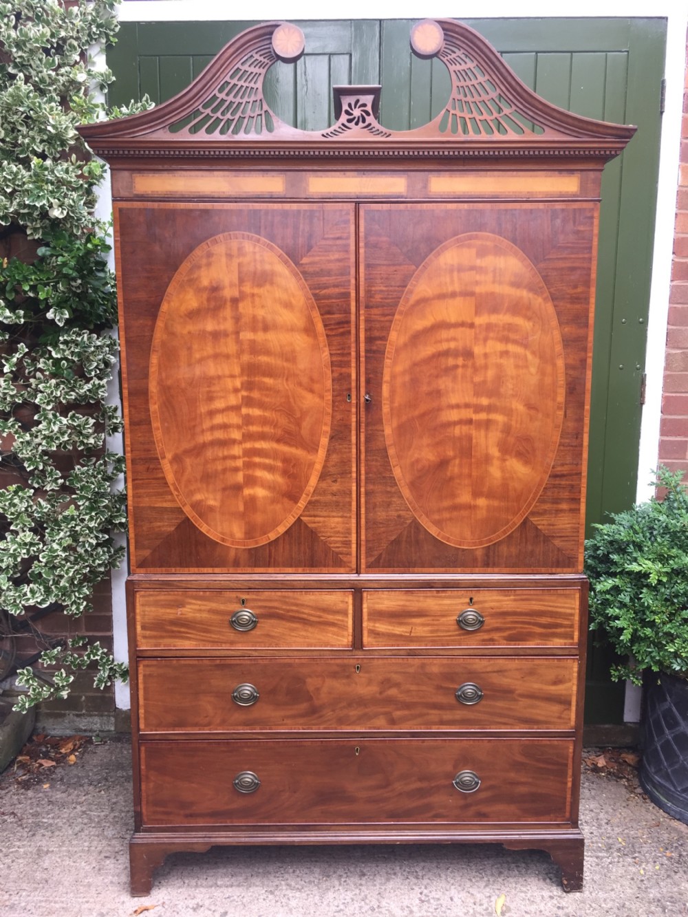 A late 18th-century George III mahogany and satinwood linen press with a swan-neck pediment, oval inlaid cupboard doors, and four lower drawers. View IX