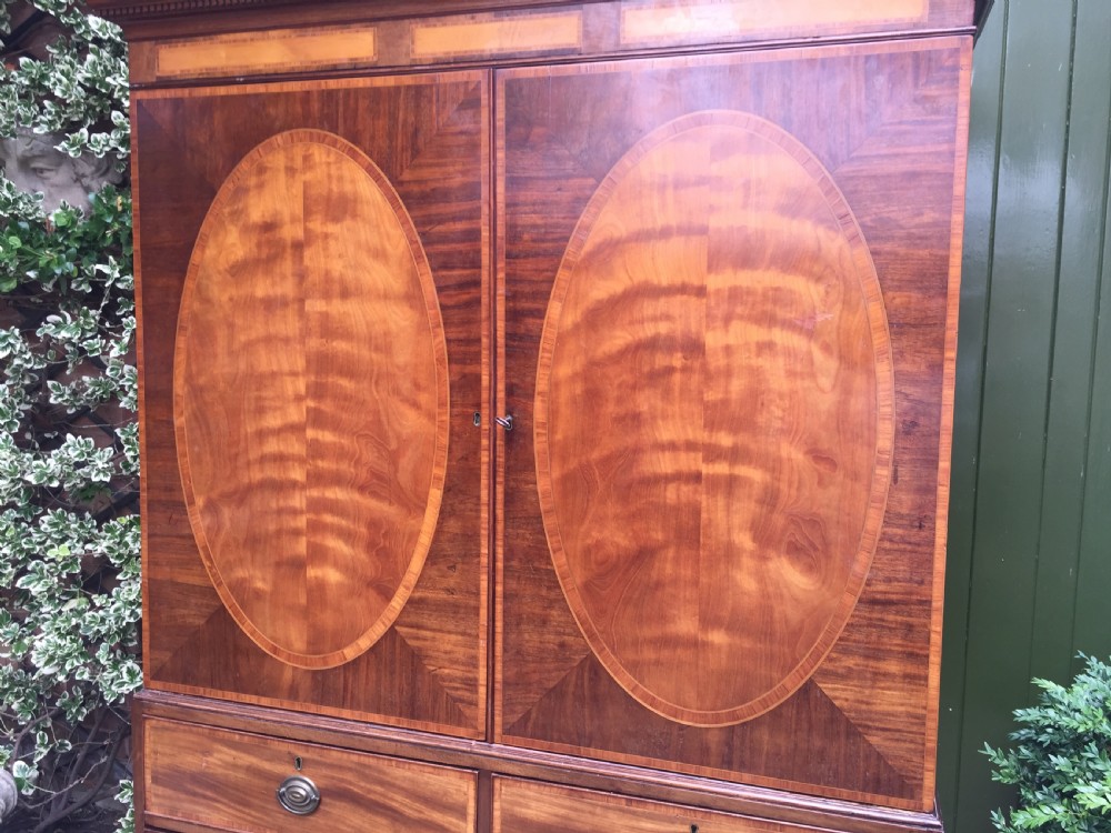 A late 18th-century George III mahogany and satinwood linen press with a swan-neck pediment, oval inlaid cupboard doors, and four lower drawers. View X