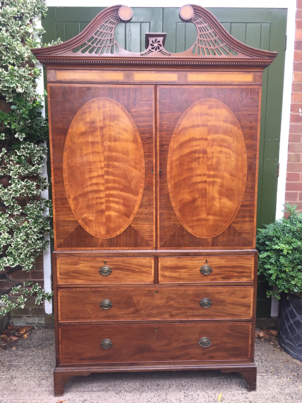 A late 18th-century George III mahogany and satinwood linen press with a swan-neck pediment, oval inlaid cupboard doors, and four lower drawers. View XX