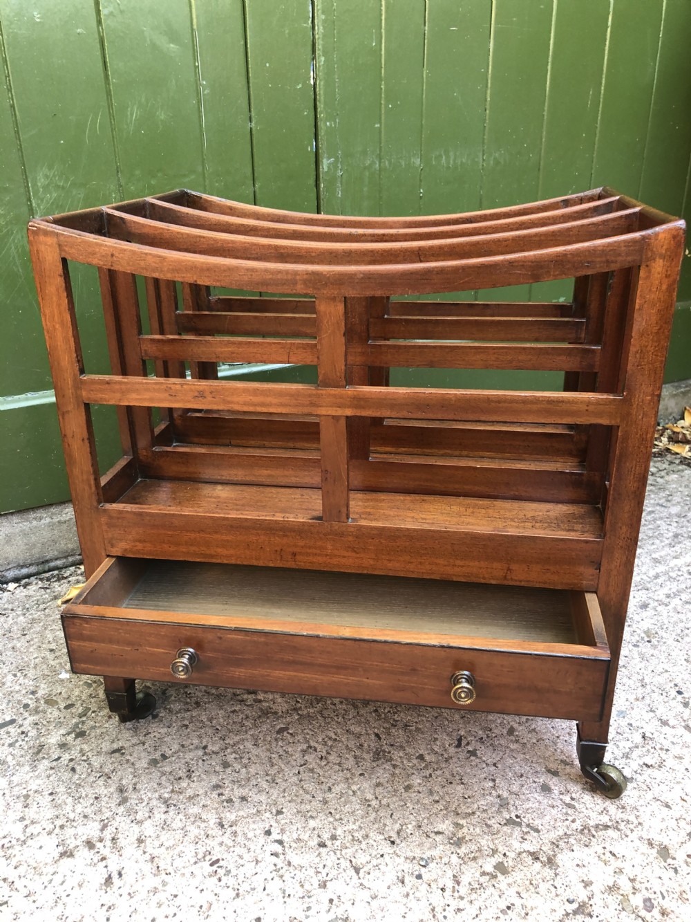 A late George III mahogany canterbury featuring slatted dividers, a concave top crest rail, a single base drawer with brass knobs, and square legs ending in brass castors. View IX
