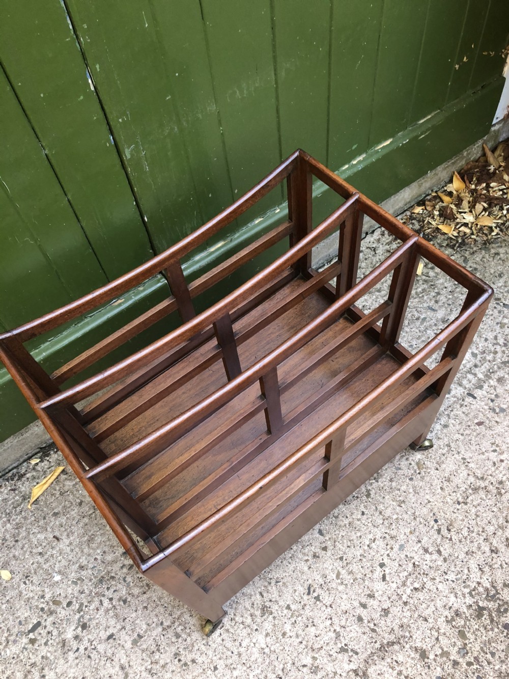 A late George III mahogany canterbury featuring slatted dividers, a concave top crest rail, a single base drawer with brass knobs, and square legs ending in brass castors. View X