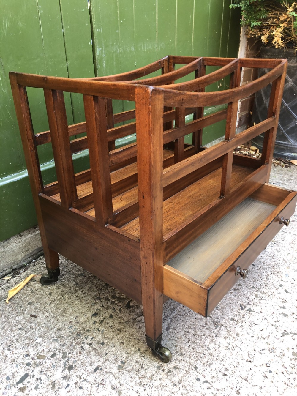 A late George III mahogany canterbury featuring slatted dividers, a concave top crest rail, a single base drawer with brass knobs, and square legs ending in brass castors. View XII
