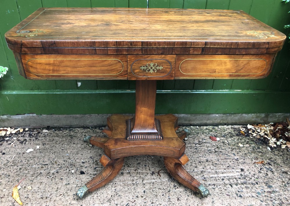 An early nineteenth-century Regency rosewood fold-over card table featuring decorative brass line inlay, a central pedestal, and splayed legs with brass claw feet.