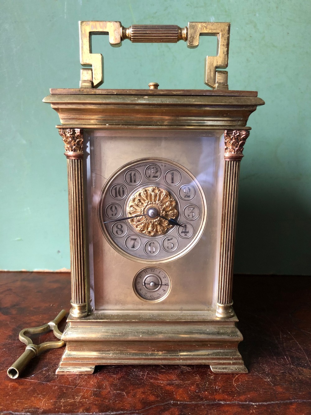 A late nineteenth-century French gilt brass carriage clock featuring fluted Corinthian corner columns, a carrying handle, a central dial with Arabic numerals, a lower subsidiary dial, and an adjacent winding key.