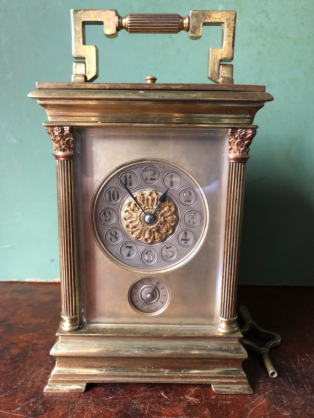 A late nineteenth-century French gilt brass carriage clock featuring fluted Corinthian corner columns, a carrying handle, a central dial with Arabic numerals, a lower subsidiary dial, and an adjacent winding key. View XVII