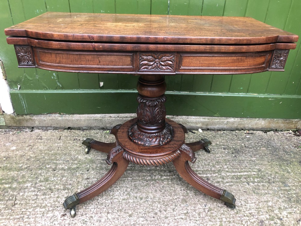 An early 19th-century Regency mahogany fold-over card table featuring carved floral frieze panels, a bulbous carved pedestal stem, and four splayed legs with brass castors.