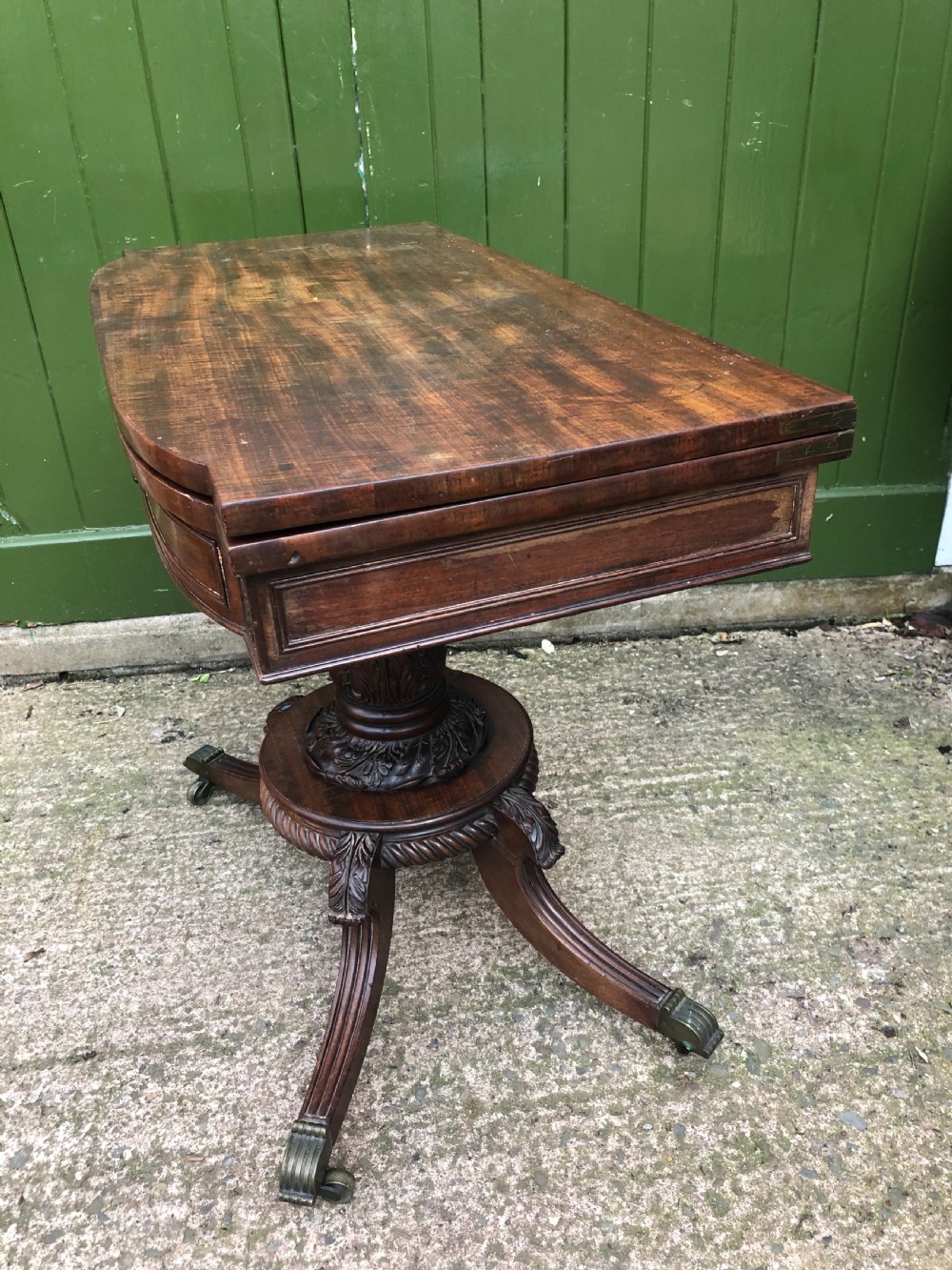 An early 19th-century Regency mahogany fold-over card table featuring carved floral frieze panels, a bulbous carved pedestal stem, and four splayed legs with brass castors. View II