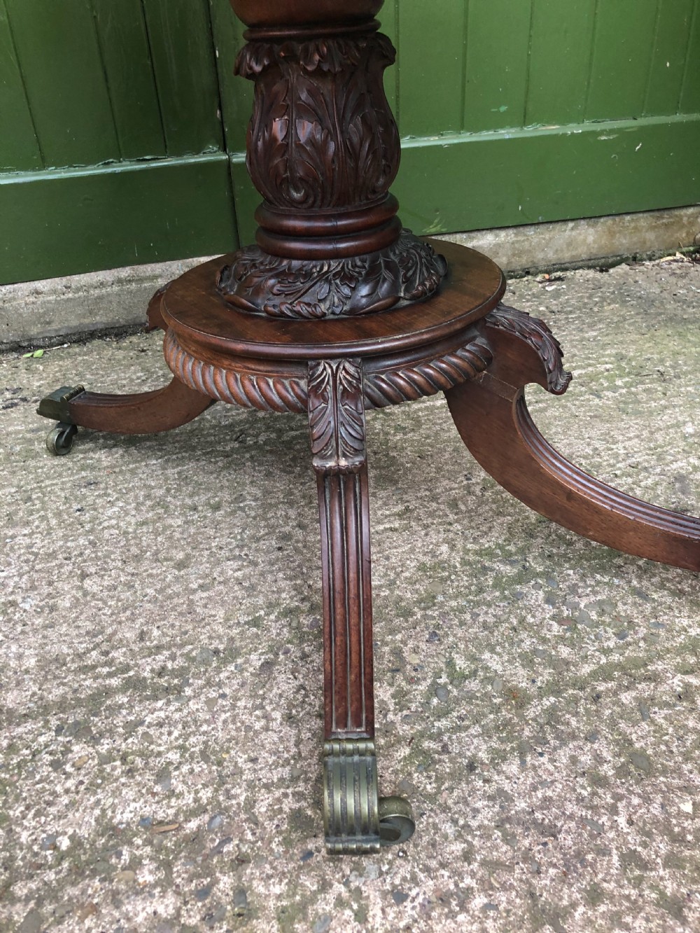 An early 19th-century Regency mahogany fold-over card table featuring carved floral frieze panels, a bulbous carved pedestal stem, and four splayed legs with brass castors. View V
