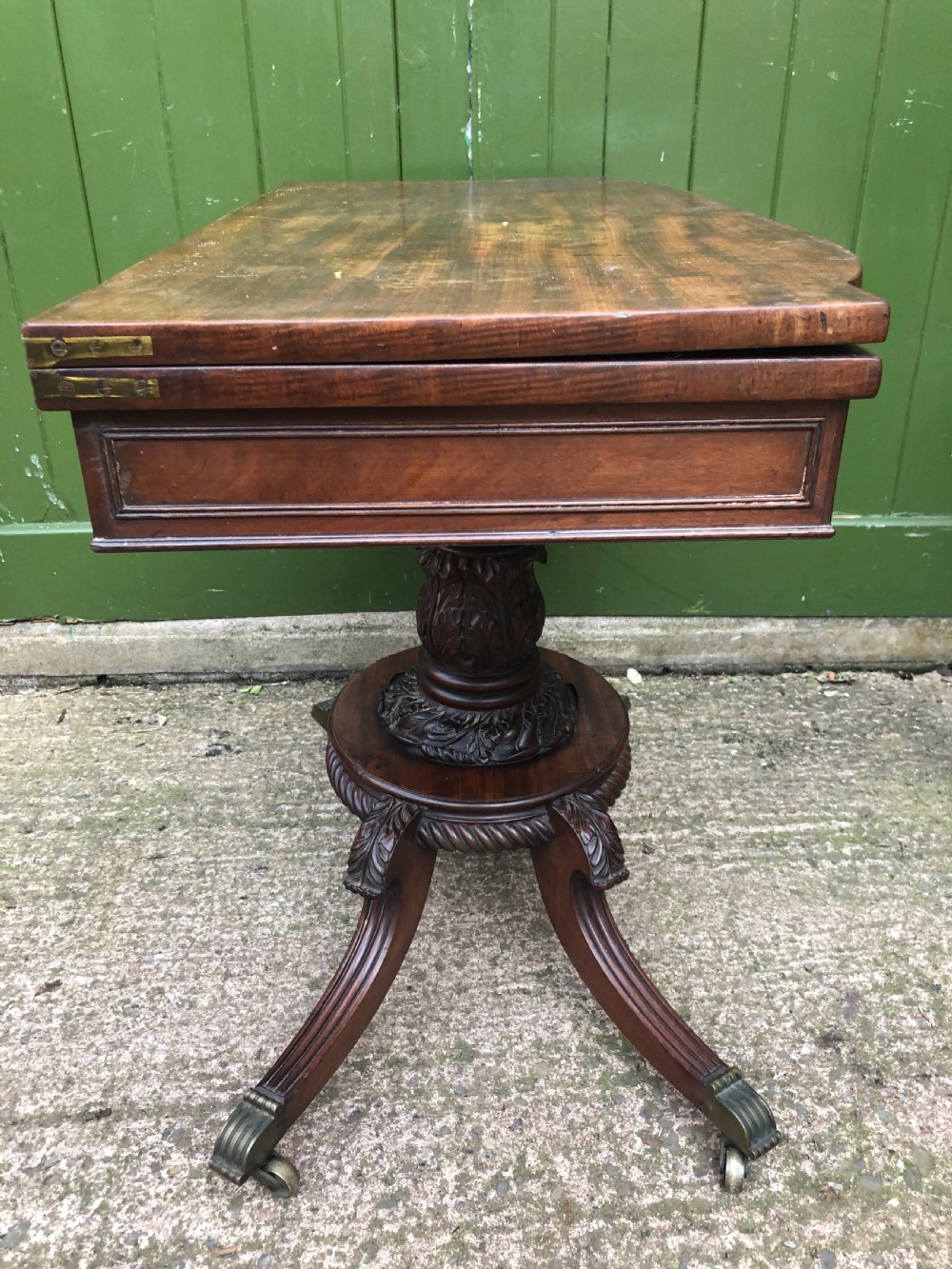 An early 19th-century Regency mahogany fold-over card table featuring carved floral frieze panels, a bulbous carved pedestal stem, and four splayed legs with brass castors. View IX