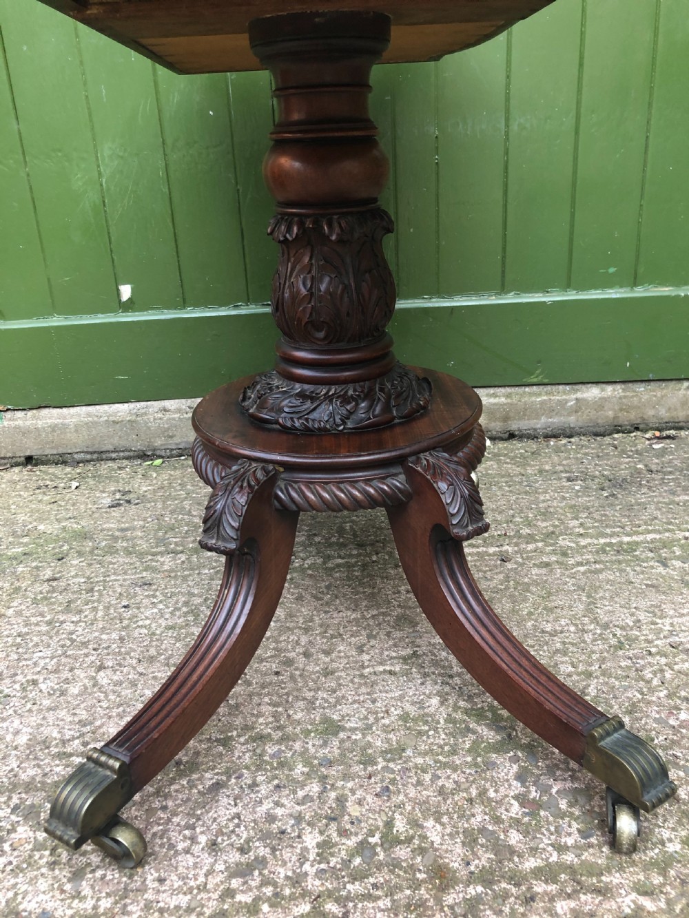 An early 19th-century Regency mahogany fold-over card table featuring carved floral frieze panels, a bulbous carved pedestal stem, and four splayed legs with brass castors. View X