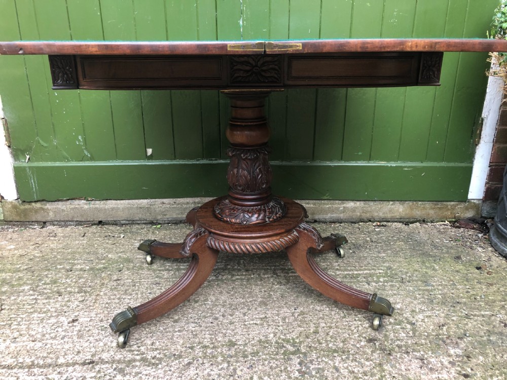 An early 19th-century Regency mahogany fold-over card table featuring carved floral frieze panels, a bulbous carved pedestal stem, and four splayed legs with brass castors. View XI