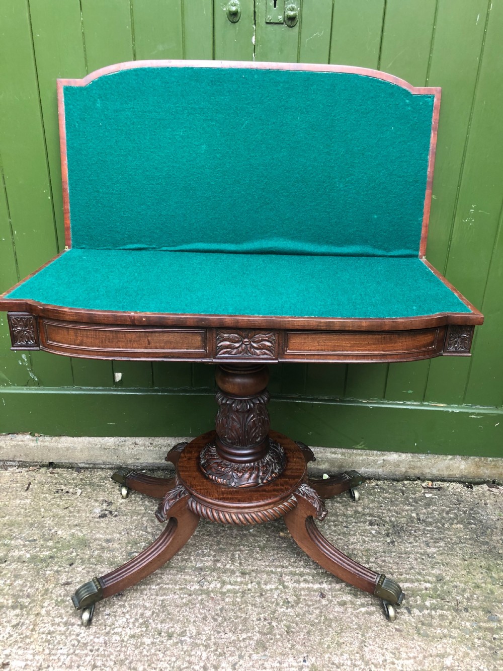 An early 19th-century Regency mahogany fold-over card table featuring carved floral frieze panels, a bulbous carved pedestal stem, and four splayed legs with brass castors. View XII