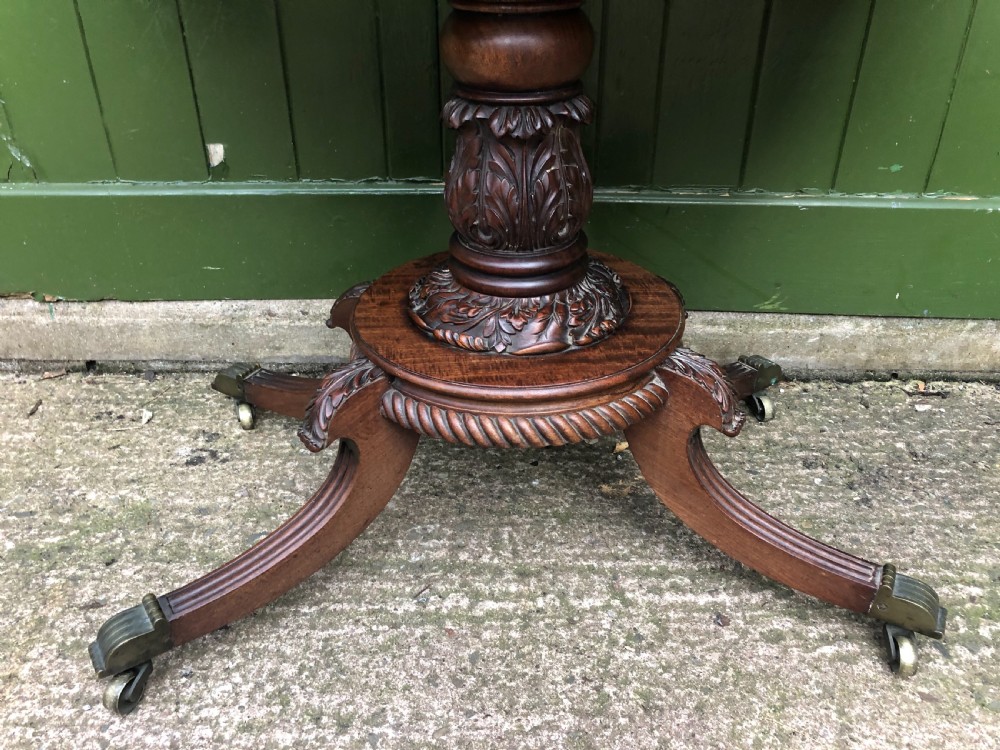 An early 19th-century Regency mahogany fold-over card table featuring carved floral frieze panels, a bulbous carved pedestal stem, and four splayed legs with brass castors. View XV