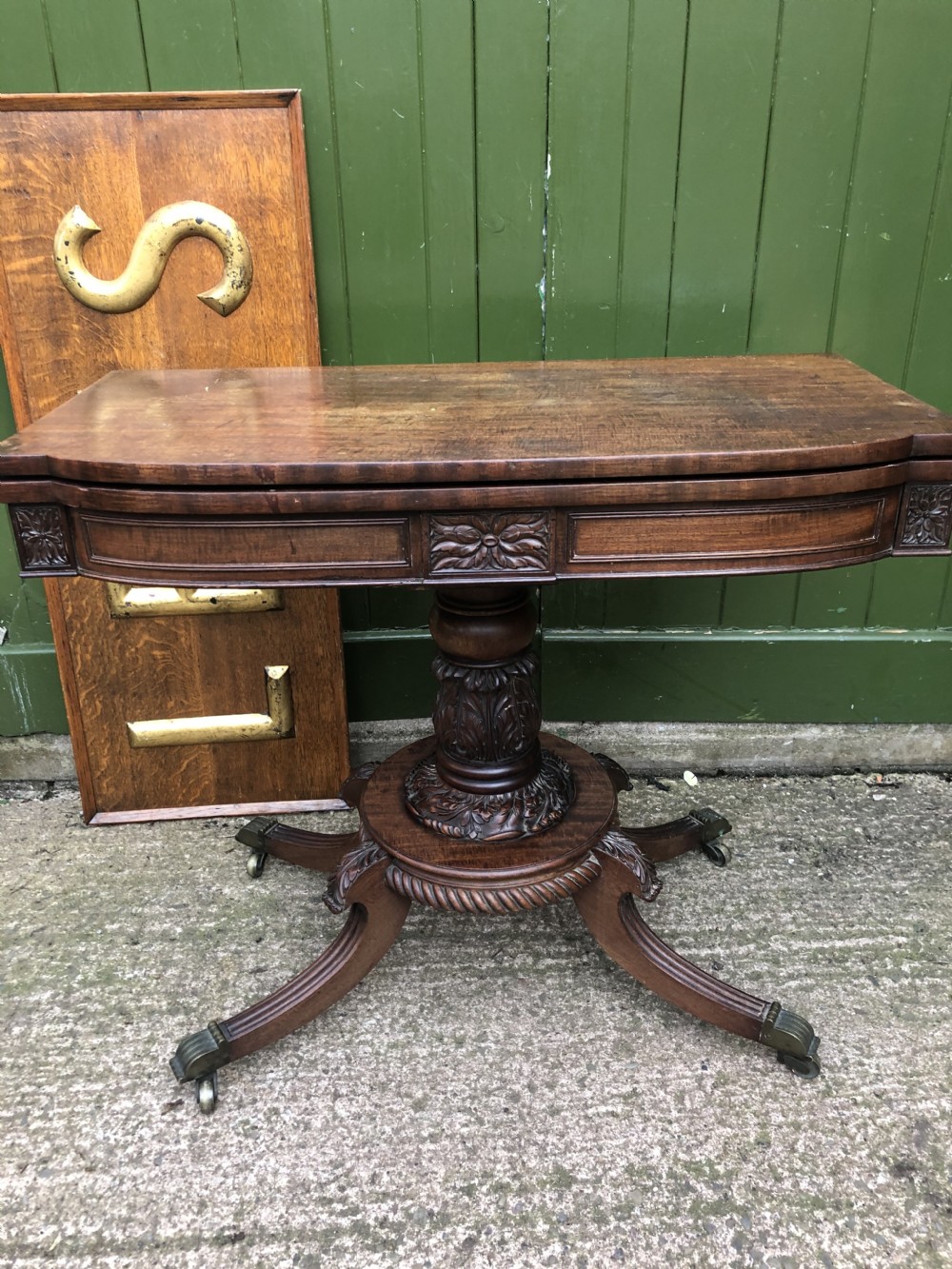 An early 19th-century Regency mahogany fold-over card table featuring carved floral frieze panels, a bulbous carved pedestal stem, and four splayed legs with brass castors. View XIX