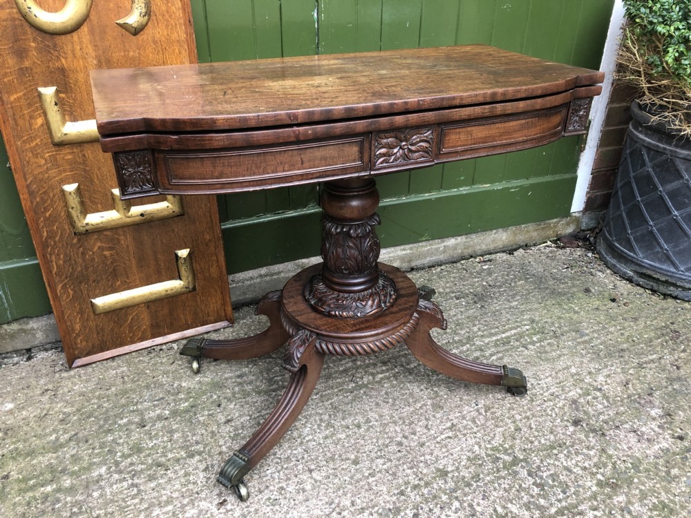 An early 19th-century Regency mahogany fold-over card table featuring carved floral frieze panels, a bulbous carved pedestal stem, and four splayed legs with brass castors. View XX