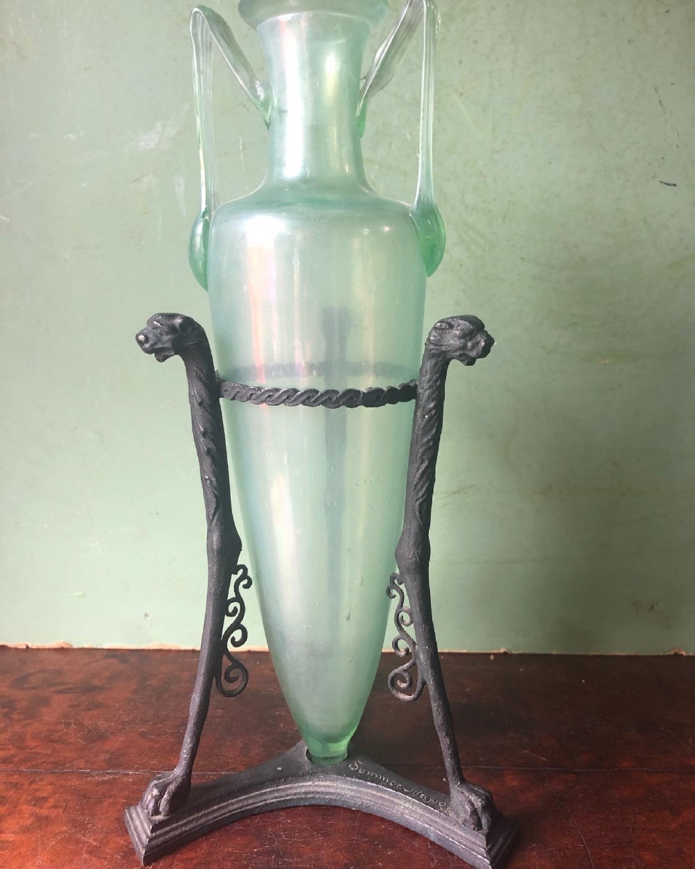A late 19th-century patinated bronze tripod stand with feline-headed legs holding a pale green glass amphora vessel.