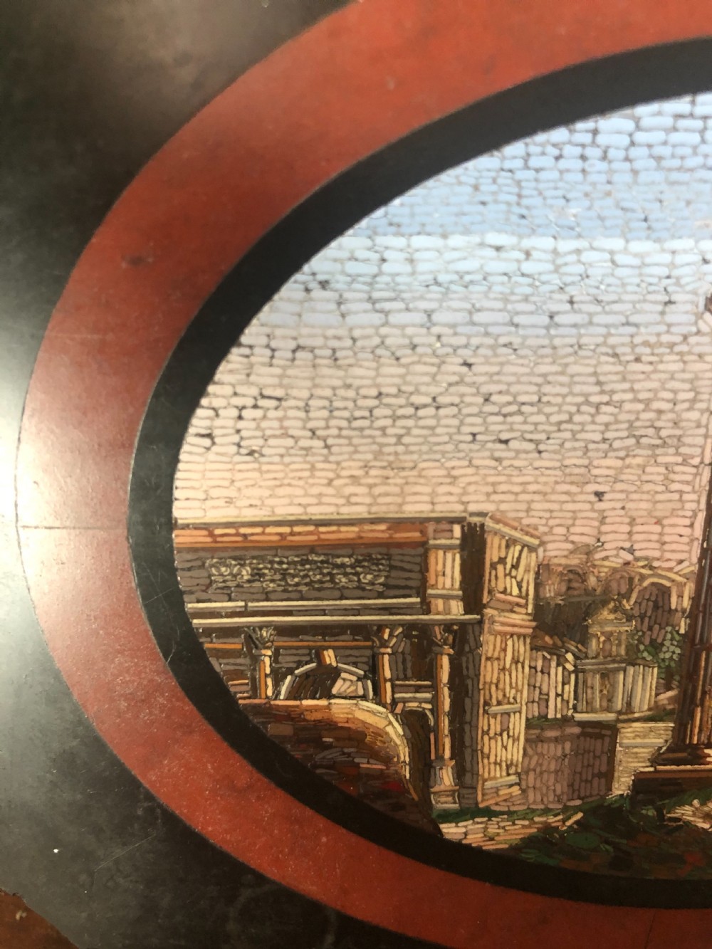 A cartouche-shaped black marble paperweight with a terracotta-coloured inlay ring surrounding a central oval micro-mosaic depicting classical Roman architectural ruins. View VI