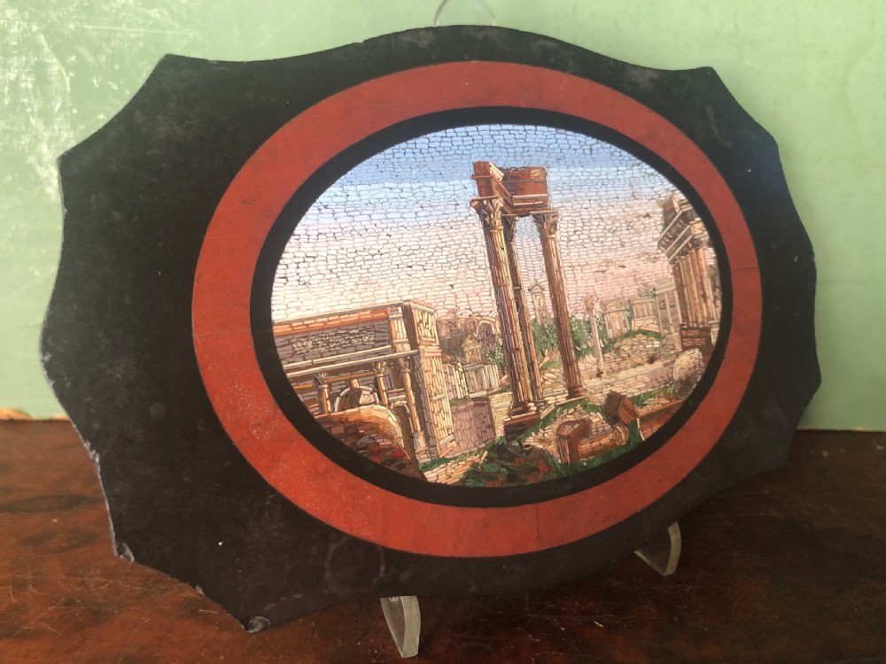A cartouche-shaped black marble paperweight with a terracotta-coloured inlay ring surrounding a central oval micro-mosaic depicting classical Roman architectural ruins. View VII