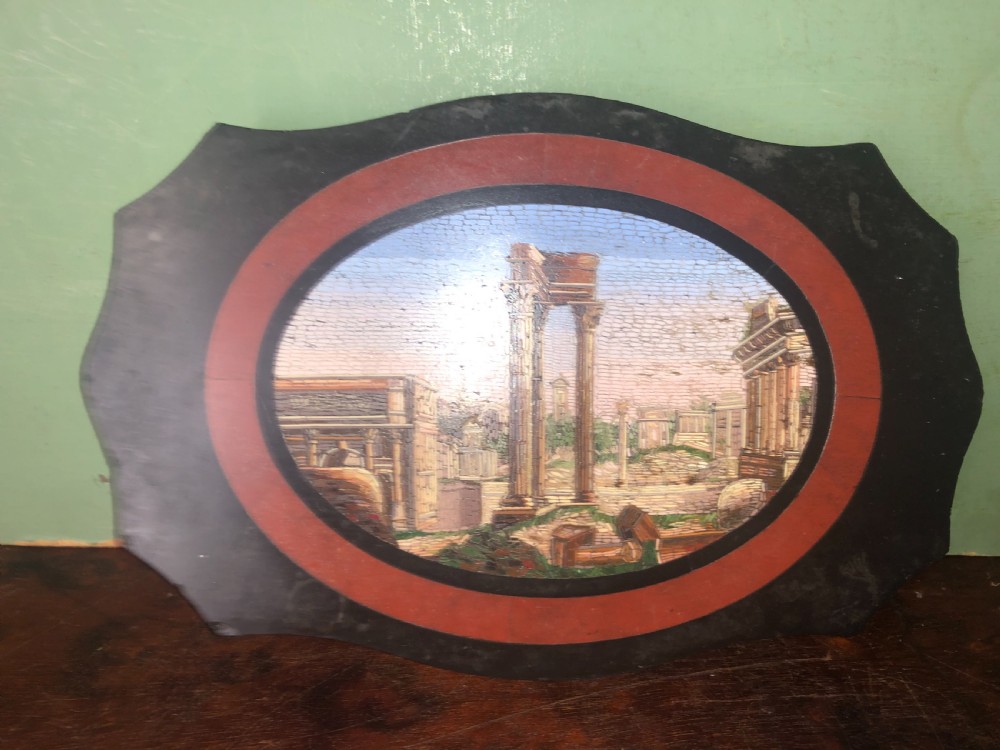 A cartouche-shaped black marble paperweight with a terracotta-coloured inlay ring surrounding a central oval micro-mosaic depicting classical Roman architectural ruins. View VIII