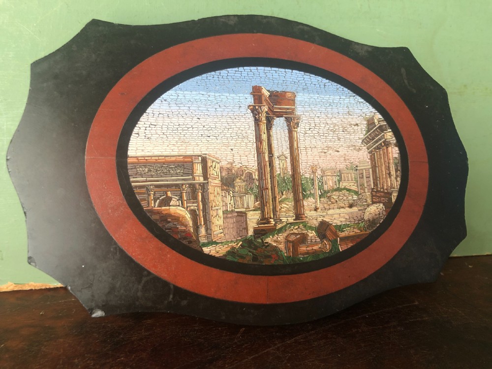 A cartouche-shaped black marble paperweight with a terracotta-coloured inlay ring surrounding a central oval micro-mosaic depicting classical Roman architectural ruins. View X