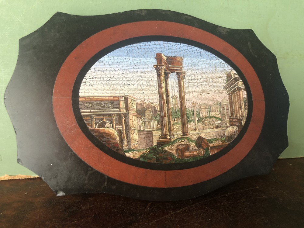 A cartouche-shaped black marble paperweight with a terracotta-coloured inlay ring surrounding a central oval micro-mosaic depicting classical Roman architectural ruins.