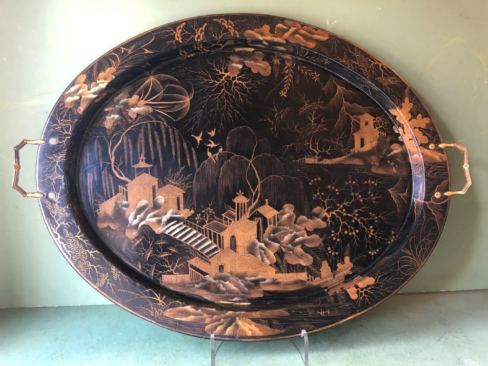 An oval black papier-mache tray featuring detailed gold chinoiserie landscape decoration and two side handles.