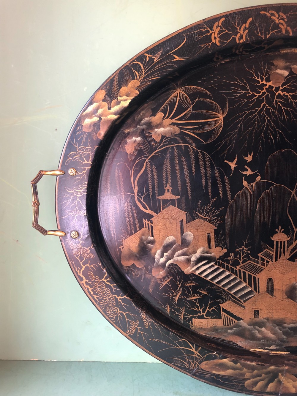 An oval black papier-mache tray featuring detailed gold chinoiserie landscape decoration and two side handles. View IV