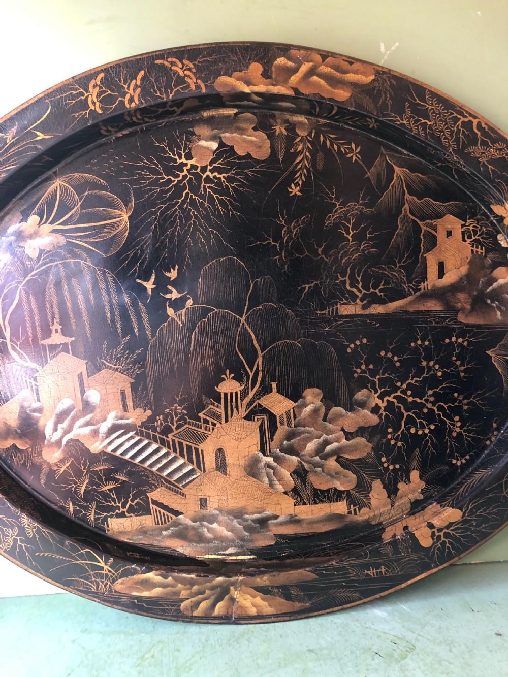 An oval black papier-mache tray featuring detailed gold chinoiserie landscape decoration and two side handles. View V
