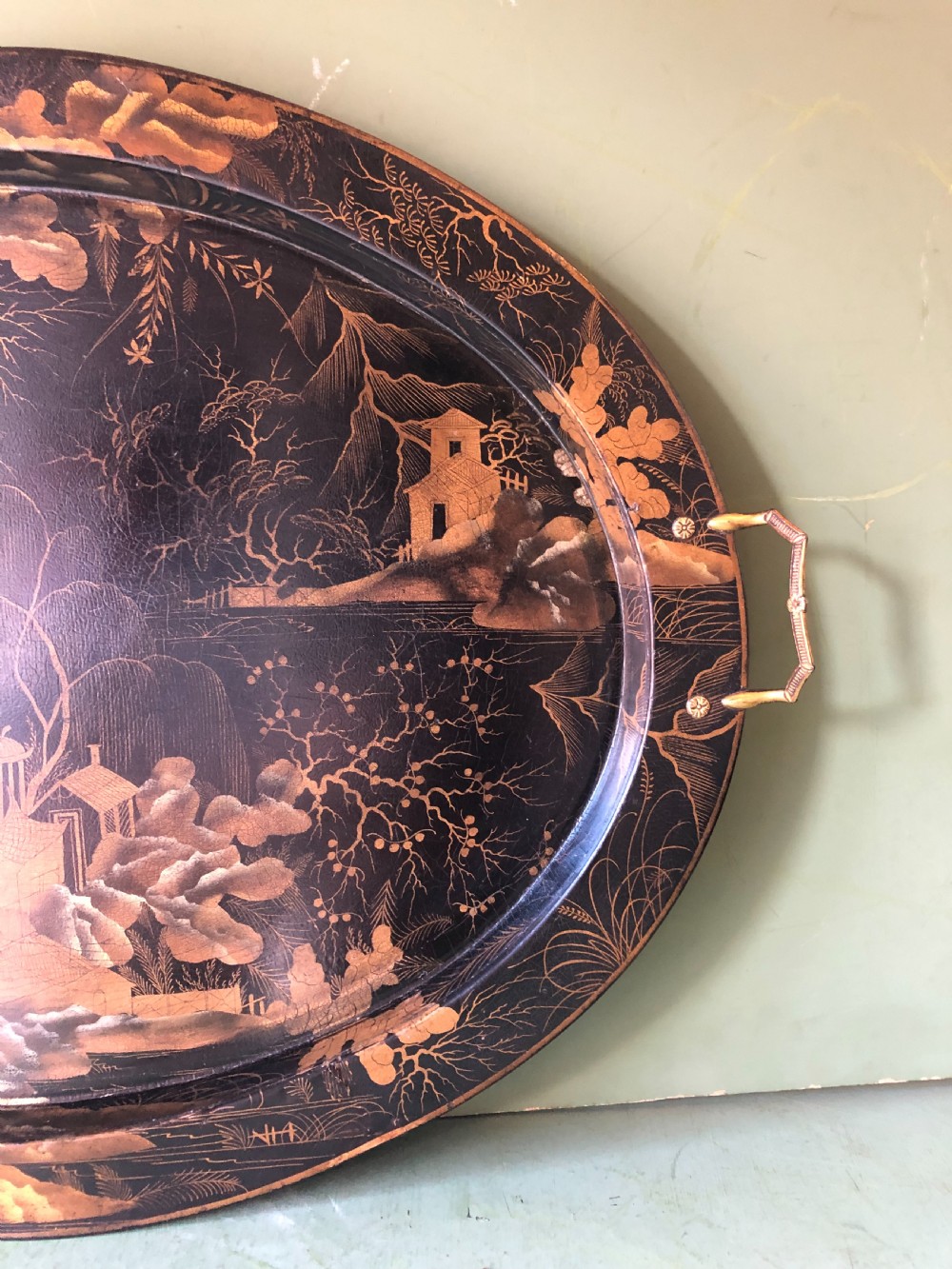 An oval black papier-mache tray featuring detailed gold chinoiserie landscape decoration and two side handles. View VII