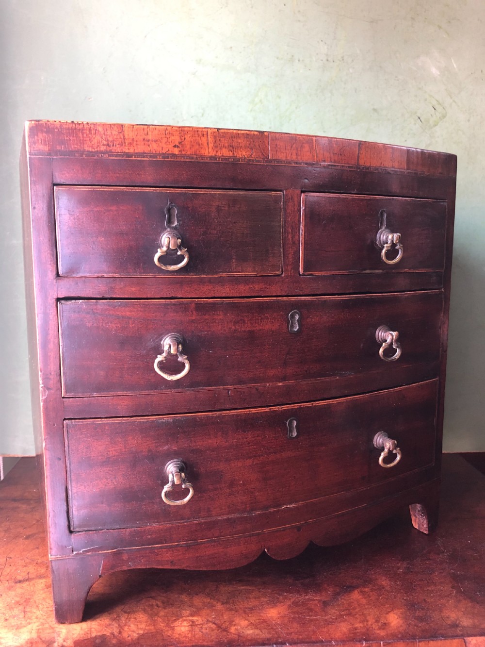 An early nineteenth-century miniature mahogany bow-front chest of drawers featuring brass ring handles and keyholes. View II
