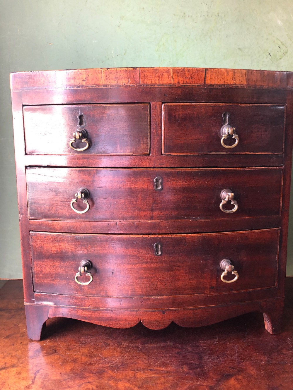 An early nineteenth-century miniature mahogany bow-front chest of drawers featuring brass ring handles and keyholes. View IV