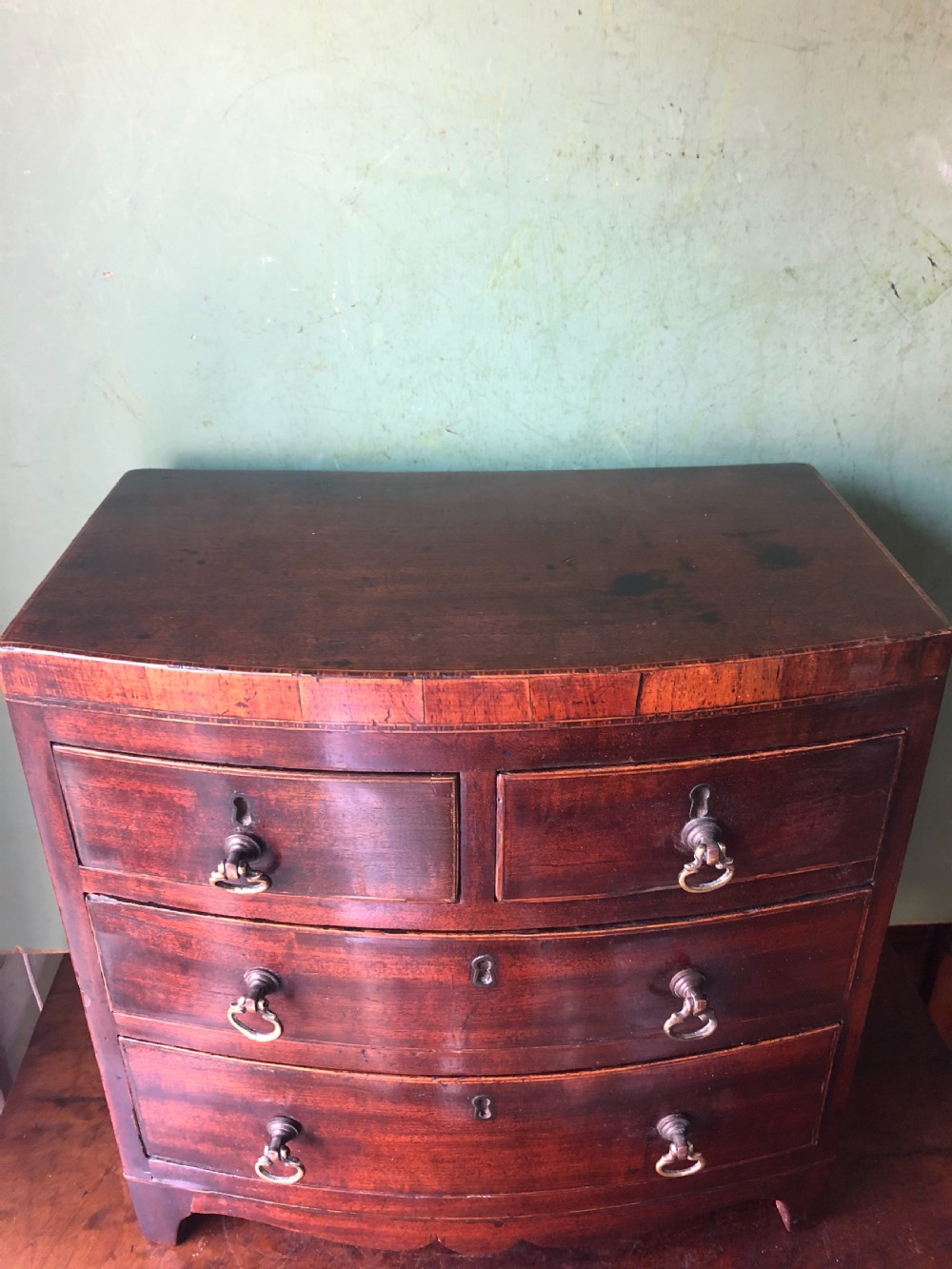 An early nineteenth-century miniature mahogany bow-front chest of drawers featuring brass ring handles and keyholes. View V