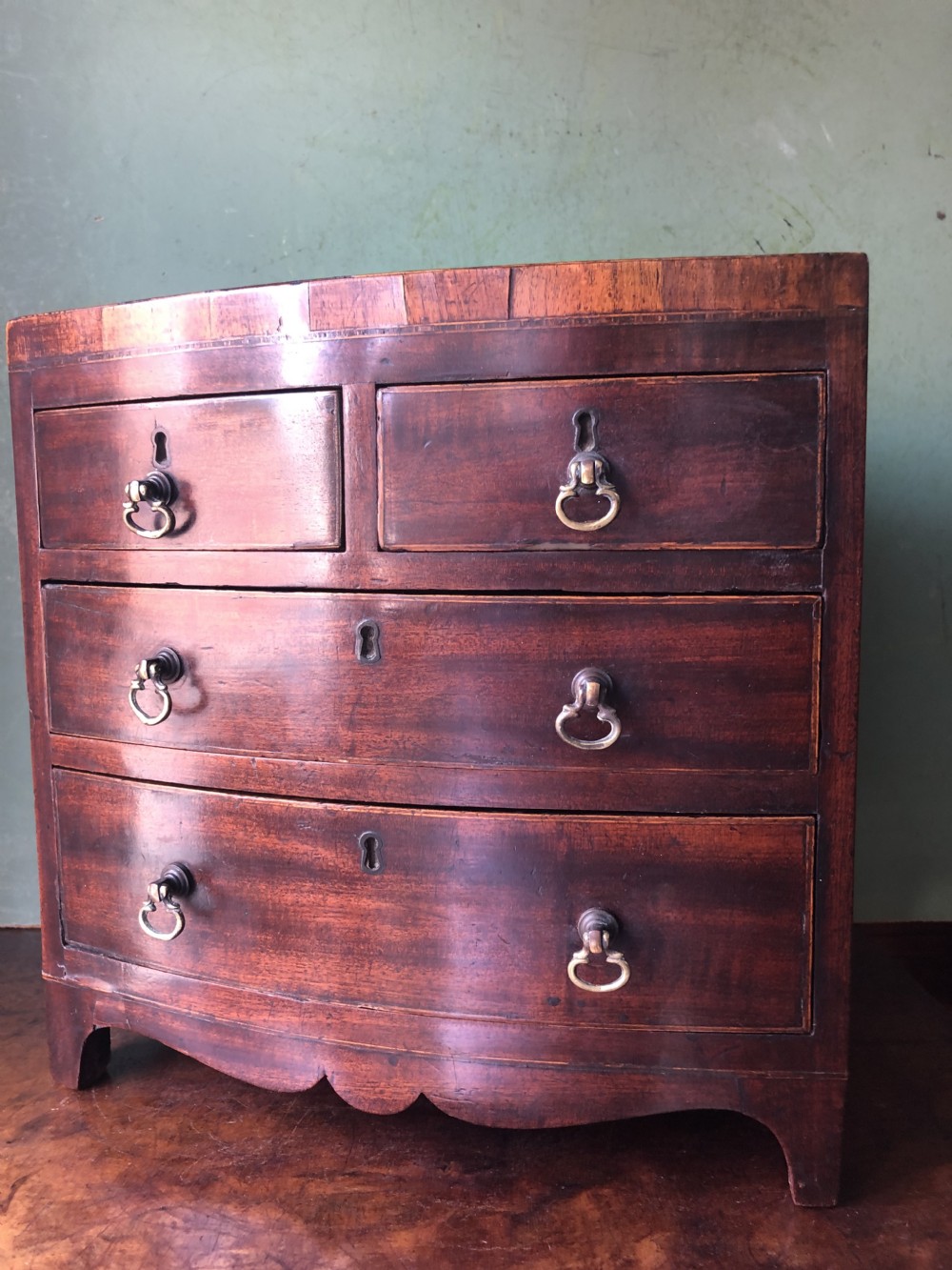 An early nineteenth-century miniature mahogany bow-front chest of drawers featuring brass ring handles and keyholes. View XIV