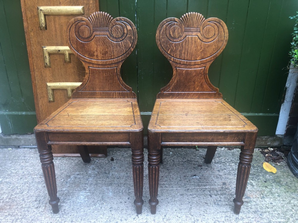 A pair of early nineteenth-century Regency oak hall chairs featuring carved scroll backs with shell motifs, solid timber seats, and reeded front legs.