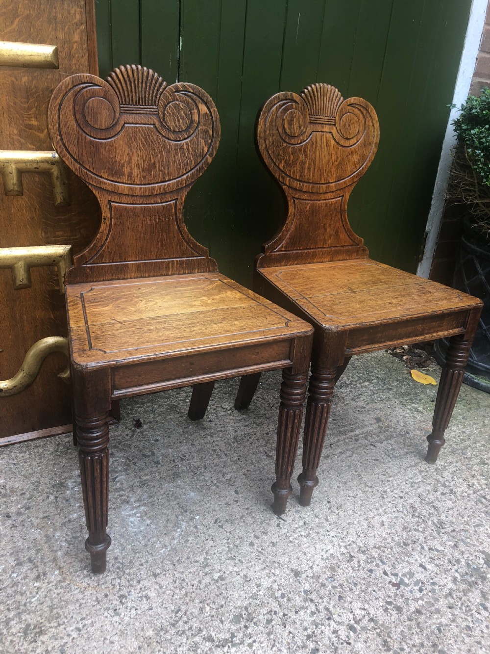 A pair of early nineteenth-century Regency oak hall chairs featuring carved scroll backs with shell motifs, solid timber seats, and reeded front legs. View II