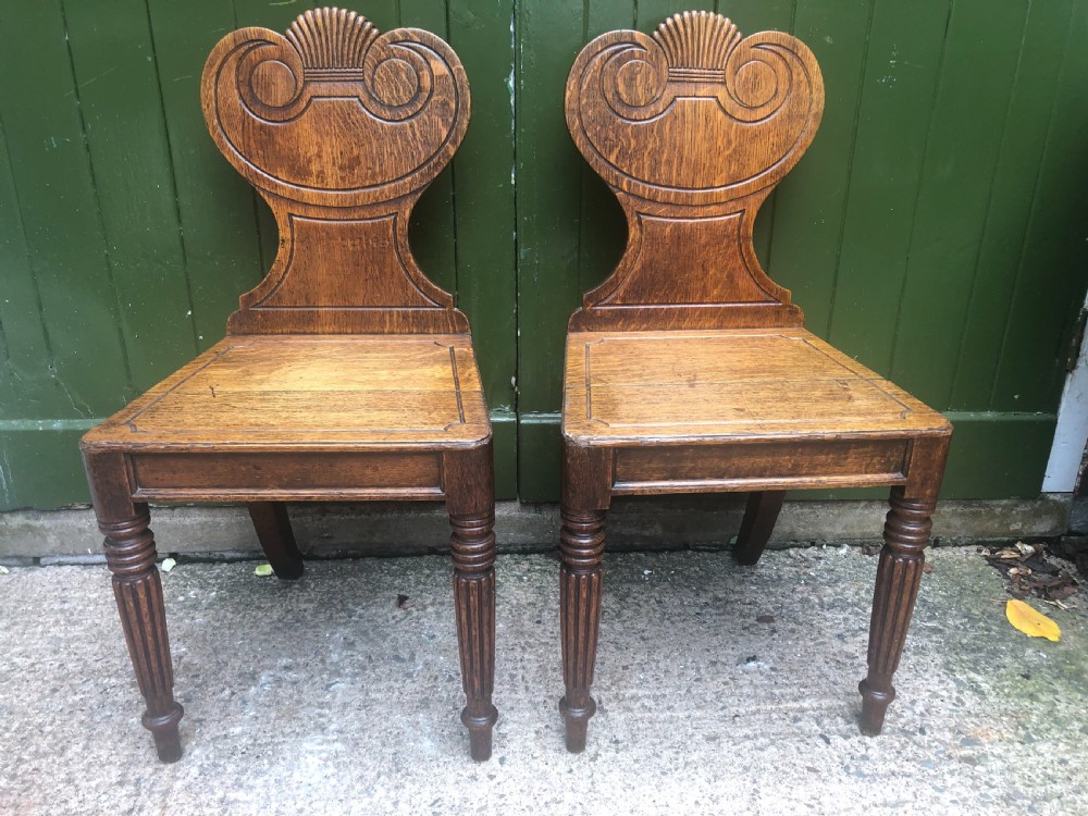 A pair of early nineteenth-century Regency oak hall chairs featuring carved scroll backs with shell motifs, solid timber seats, and reeded front legs. View V