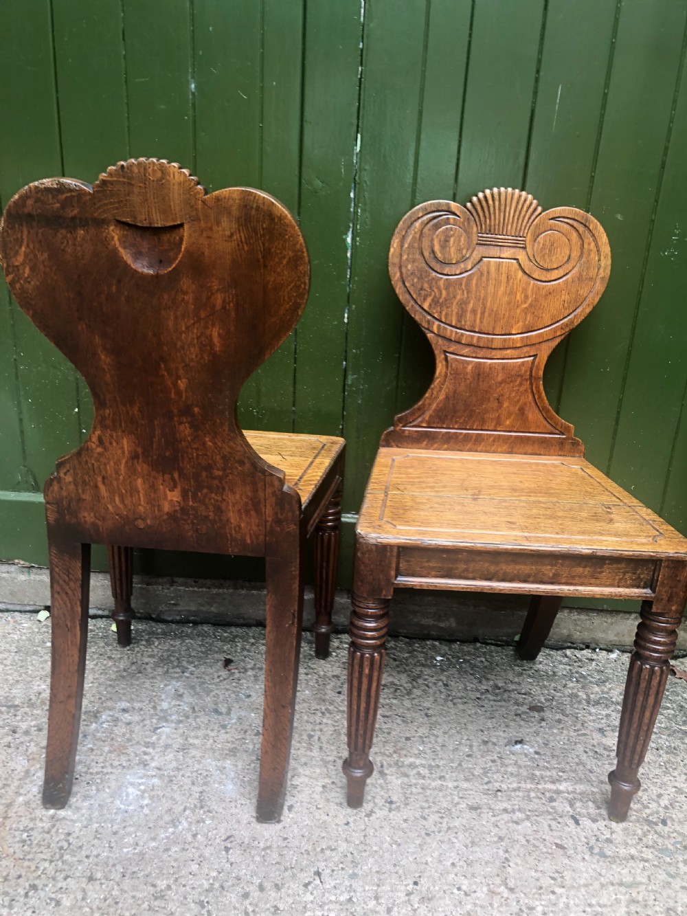 A pair of early nineteenth-century Regency oak hall chairs featuring carved scroll backs with shell motifs, solid timber seats, and reeded front legs. View X