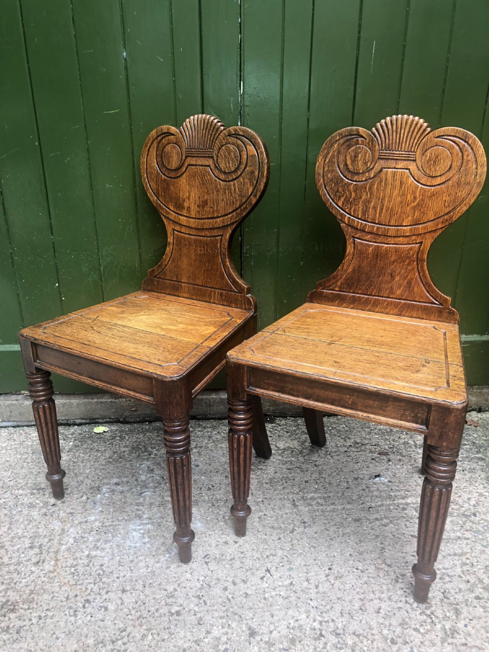 A pair of early nineteenth-century Regency oak hall chairs featuring carved scroll backs with shell motifs, solid timber seats, and reeded front legs. View XIII