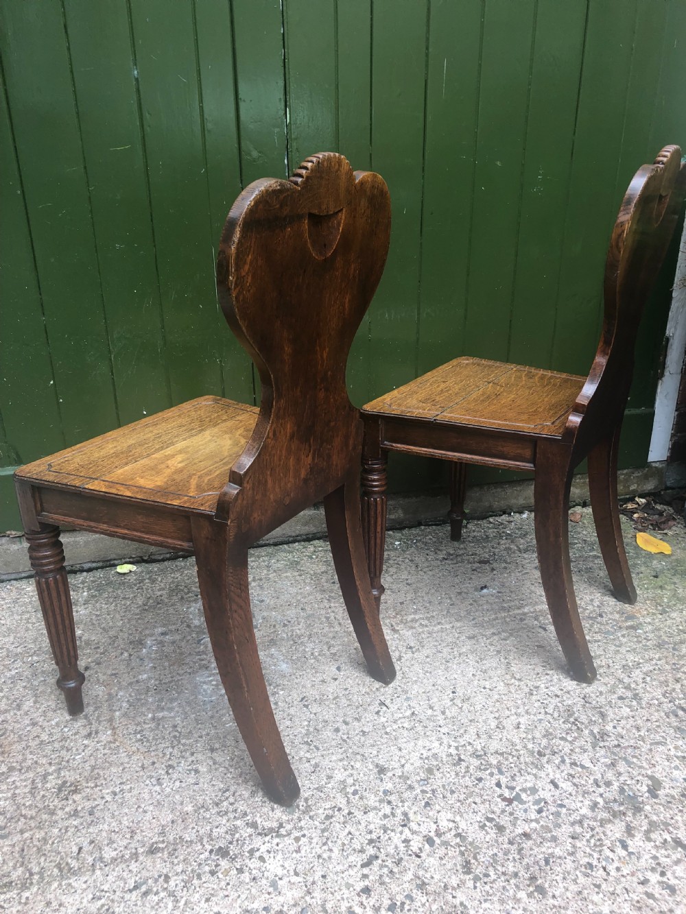 A pair of early nineteenth-century Regency oak hall chairs featuring carved scroll backs with shell motifs, solid timber seats, and reeded front legs. View XVII