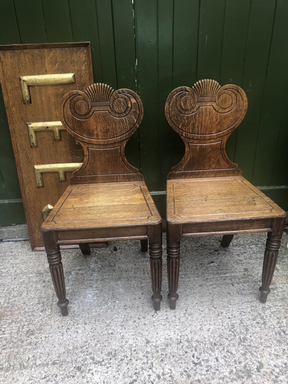 A pair of early nineteenth-century Regency oak hall chairs featuring carved scroll backs with shell motifs, solid timber seats, and reeded front legs. View XX