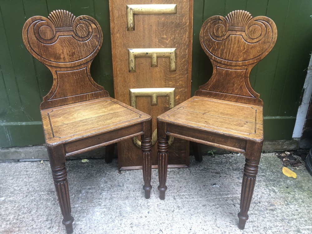 A pair of early nineteenth-century Regency oak hall chairs featuring carved scroll backs with shell motifs, solid timber seats, and reeded front legs. View XXI
