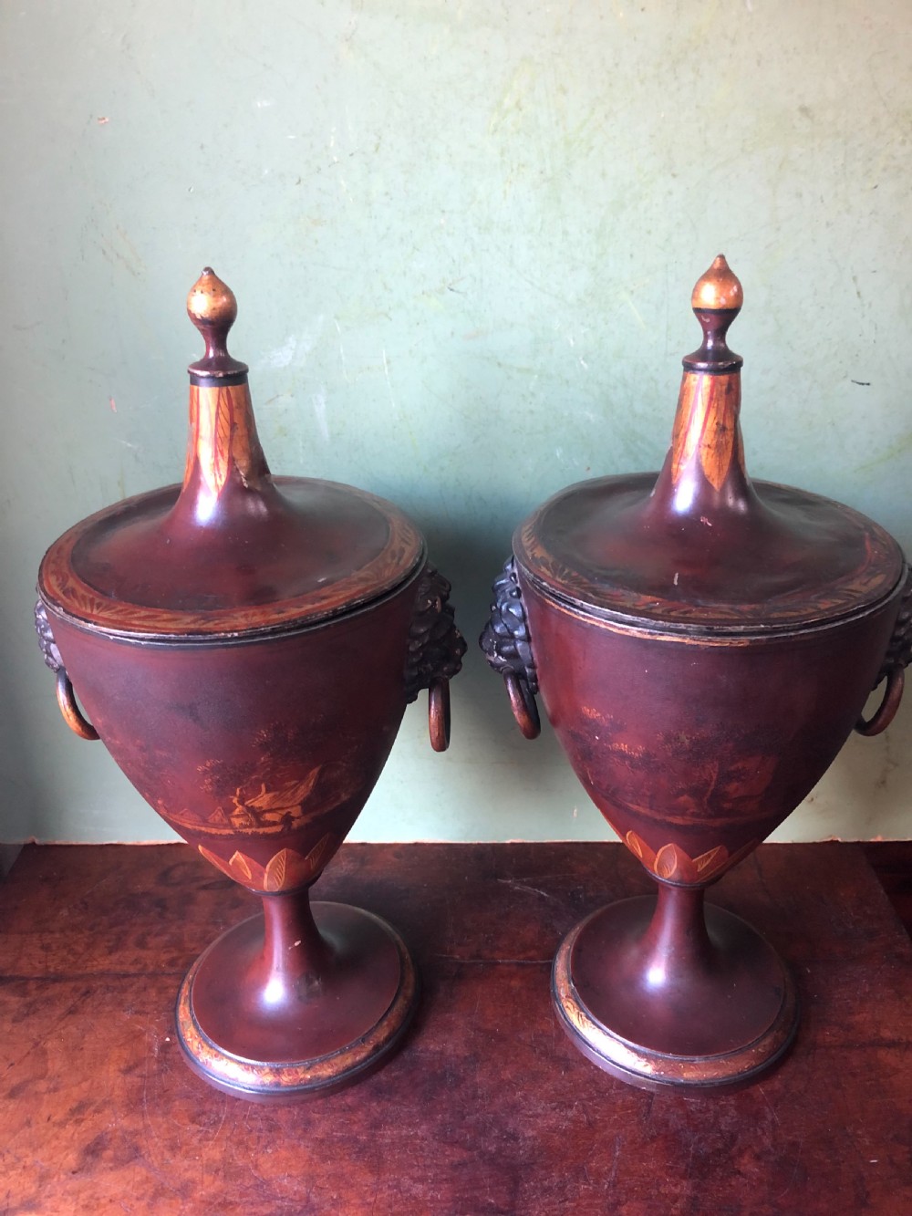 A pair of early 19th-century Dutch neoclassical chestnut urns in dark brown japanned pewter, painted with landscape scenes, leaf motifs, side ring handles, and tall finial lids. View VI