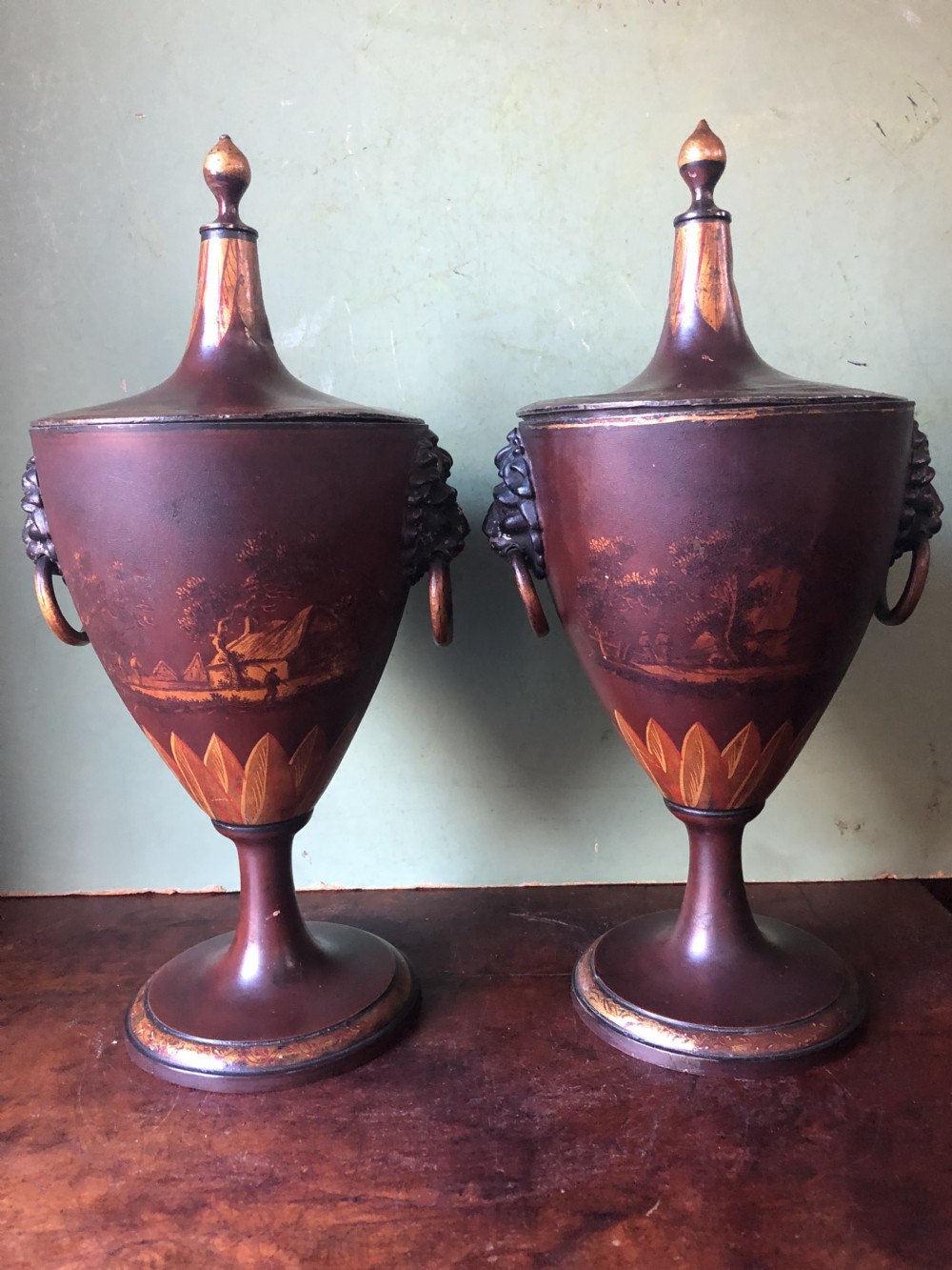 A pair of early 19th-century Dutch neoclassical chestnut urns in dark brown japanned pewter, painted with landscape scenes, leaf motifs, side ring handles, and tall finial lids.