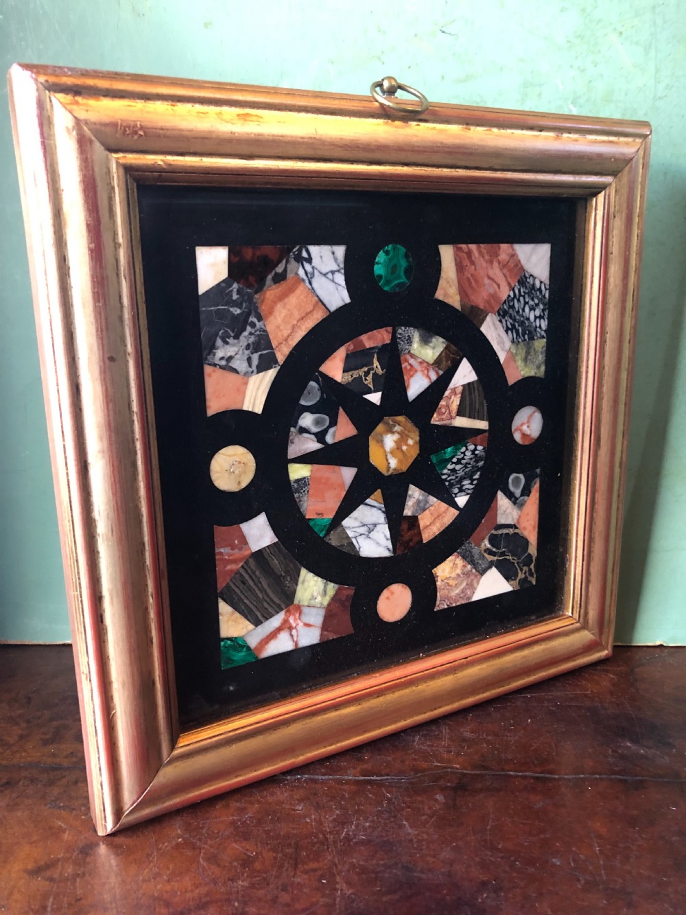 A square 19th-century Ashford black marble panel inlaid with colourful specimen marble geometric shapes forming a central star motif, set within a gilt wood frame with a top brass hanging ring. View XII