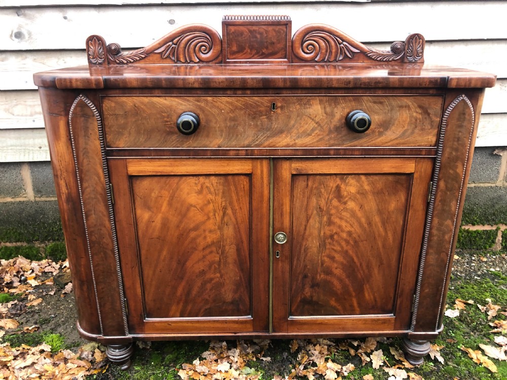 An early 19th-century William IV mahogany serving buffet featuring a carved scroll upstand, a top drawer with turned knobs, and twin panelled cupboard doors.