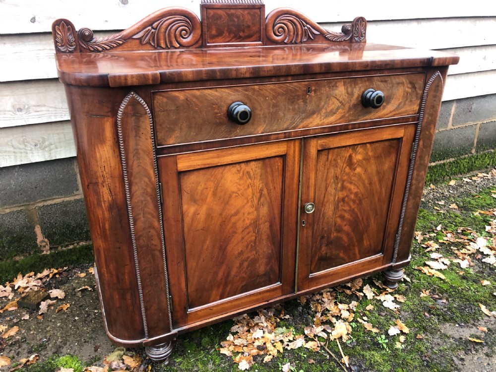 An early 19th-century William IV mahogany serving buffet featuring a carved scroll upstand, a top drawer with turned knobs, and twin panelled cupboard doors. View VI