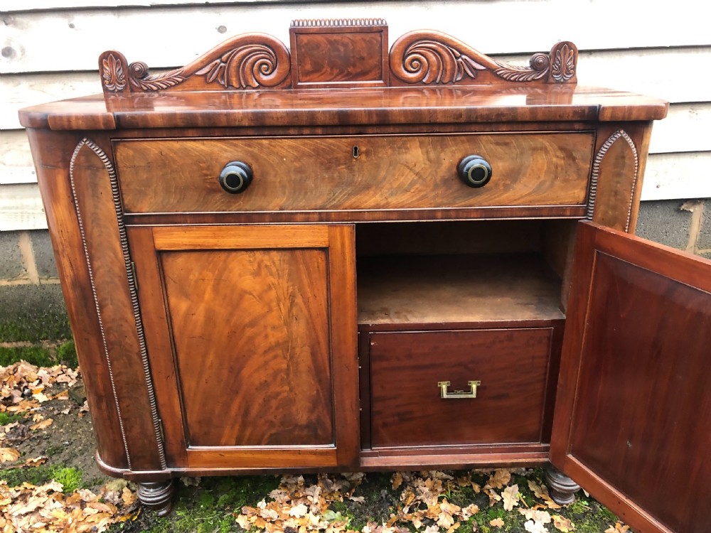 An early 19th-century William IV mahogany serving buffet featuring a carved scroll upstand, a top drawer with turned knobs, and twin panelled cupboard doors. View XI