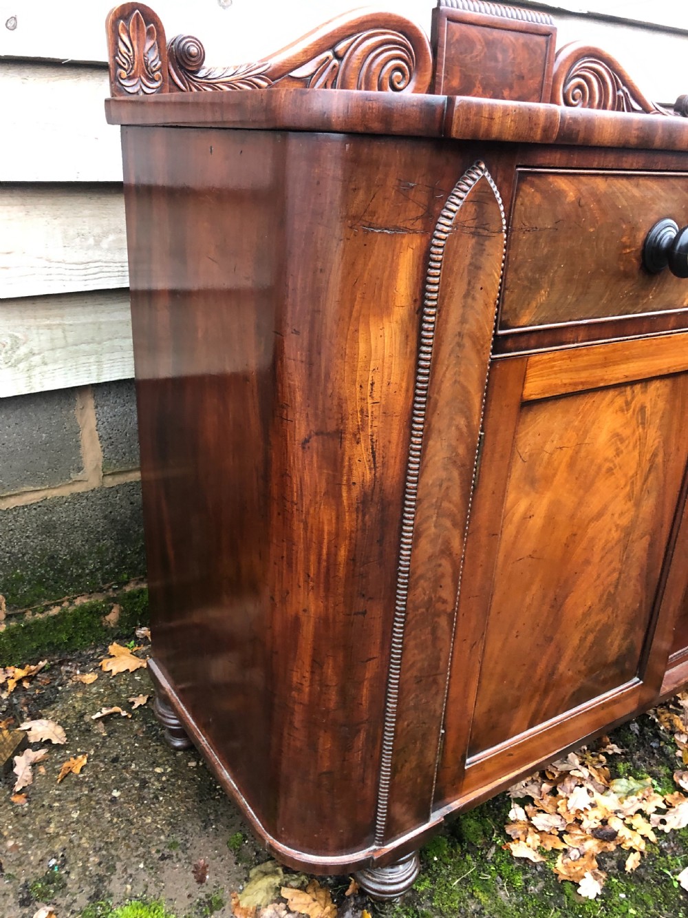 An early 19th-century William IV mahogany serving buffet featuring a carved scroll upstand, a top drawer with turned knobs, and twin panelled cupboard doors. View XII