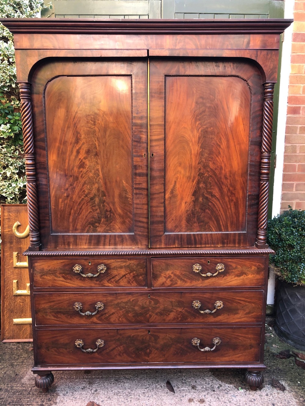 A William IV flame mahogany linen press featuring a double door upper cupboard with rope-twist side columns set above a lower chest of drawers with brass handles.