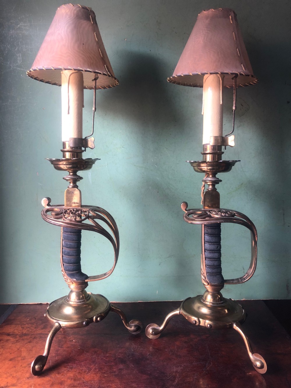 A pair of late nineteenth-century gilded brass candlelamps modelled as cavalry dress swords with basket guards, standing on tripod feet with conical shades.